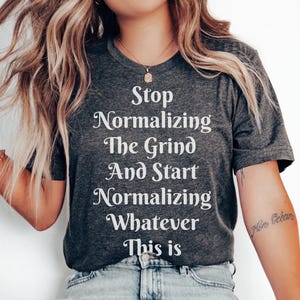 Könnte beinhalten: Ein dunkelgraues T-Shirt mit weißem Text, der "Stop Normalizing The Grind And Start Normalizing Whatever This Is" lautet.