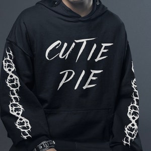 Könnte beinhalten: Schwarzer Hoodie mit weißem Text "CUTIE PIE" und weißen kettenartigen Designs an den Ärmeln.