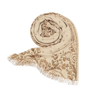 May include: A cream-colored scarf featuring a brown floral and paisley print. The fabric is arranged in a circular fold, showcasing a detailed botanical pattern with intricate line work and a decorative border, finished with a fringed edge.