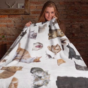 Op de afbeelding: Een witte deken met verschillende kattenillustraties in bruin, grijs en zwart. De deken toont verschillende kattenposes en -uitdrukkingen, waardoor een speels en gezellig ontwerp ontstaat. De deken wordt door een persoon vastgehouden.