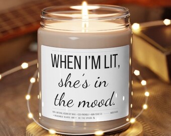 May include: A brown glass candle jar with a white label that reads "WHEN I'M LIT, she's in the mood." The candle is lit and the flame is visible. The candle is surrounded by white string lights.