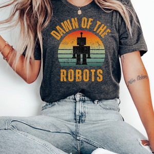 May include: A dark grey t-shirt with a vintage-style sunset graphic and the text "Dawn of the Robots". The graphic features a robot silhouette in the center of the sunset.