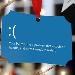 Puede incluir: Un adorno navideño azul con forma de pantalla de ordenador con un emoticono de cara triste y el texto "Su PC ha encontrado un problema que no pudo solucionar, y ahora necesita reiniciarse."