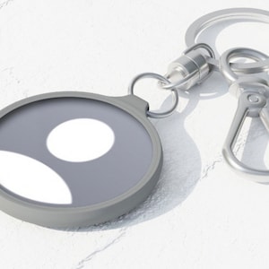 May include: A silver key chain with a round, gray, silicone charm featuring a white circle and a white crescent shape.