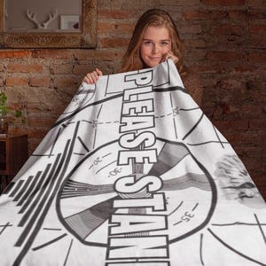 May include: A white blanket with a black and gray graphic design, featuring the text "PLEASE STAND BY" in large, bold letters. The design includes a circular pattern with lines and numbers, resembling a television test pattern.