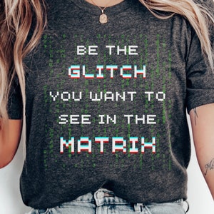 May include: A dark grey t-shirt with a white and blue pixelated graphic that says "BE THE GLITCH YOU WANT TO SEE IN THE MATRIX". The graphic is surrounded by green binary code.