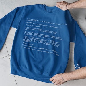 Op de afbeelding: Een blauwe sweatshirt met witte tekst die lijkt op een computerfoutmelding. De tekst bevat zinnen als "Er is een probleem gedetecteerd" en "stopfout". De sweatshirt heeft een effen blauwe kleur.