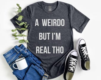 Puede incluir: Una camiseta gris oscuro con el texto blanco "A Weirdo But I'm Real Tho". La imagen incluye vaqueros azules, una taza blanca y zapatillas negras y blancas. También hay una ramita de vegetación.