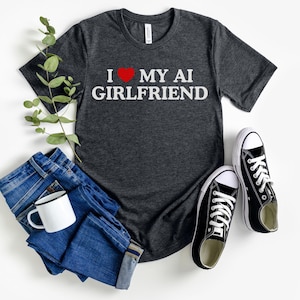 Peut inclure: T-shirt gris foncé avec l'inscription blanche "I ❤️ MY AI GIRLFRIEND" et un cœur rouge. L'image comprend également un jean bleu, une tasse blanche et des baskets noires.