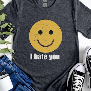 Könnte beinhalten: Dunkelgraues T-Shirt mit einem vergilbten gelben Smiley und dem weißen Text "I hate you". Das Bild zeigt blaue Jeans, einen weißen Becher und schwarz-weiße Turnschuhe.