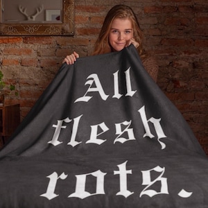 May include: A dark gray blanket with the words "All flesh rots" in white gothic lettering. The blanket is held up by a person, with a brick wall in the background.