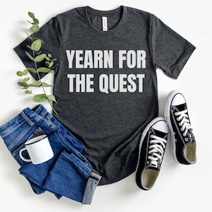 Puede incluir: Una camiseta gris oscuro con el texto "YEARN FOR THE QUEST" impreso en blanco en la parte delantera. La camiseta está colocada sobre una superficie blanca con un par de zapatillas negras y un par de pantalones vaqueros azules.