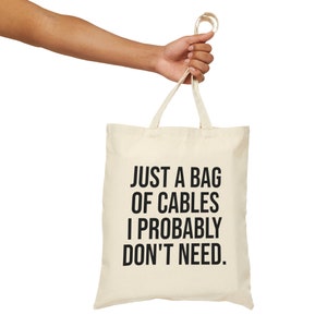 May include: A white canvas tote bag with black text that reads "Just a bag of cables I probably don't need."