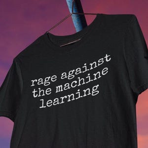 Könnte beinhalten: Schwarzes T-Shirt mit dem weißen Text "rage against the machine learning". Das Shirt hängt an einem braunen Kleiderbügel vor einem farbenfrohen Sonnenuntergangshintergrund. Das T-Shirt ist aus einem weichen Material.