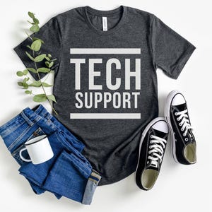 May include: Dark gray t-shirt with the words "TECH SUPPORT" in white block letters. The shirt is paired with blue jeans, a white mug, and black canvas sneakers with white laces. A sprig of greenery adds a decorative touch.
