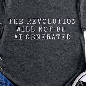Puede incluir: Camiseta gris oscuro con el texto blanco "THE REVOLUTION WILL NOT BE AI GENERATED". La camiseta se combina con vaqueros azules y zapatillas deportivas blancas y negras.