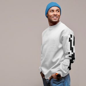 Peut inclure: Un sweat-shirt blanc avec un motif pixelisé noir sur la manche droite. La personne porte un bonnet bleu.