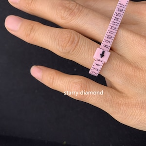 May include: A pink measuring tape with black numbers is wrapped around a finger to measure ring size. The tape reads "8 9 10 11 12 13 14 15 16". The text "starry diamond" is visible below the tape.