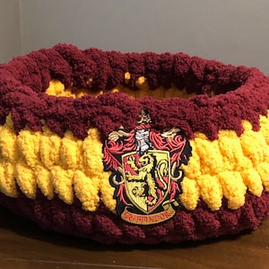 Peut inclure: Un lit pour animal de compagnie en crochet de couleur marron et jaune avec un blason de la maison Gryffondor brodé sur le devant.