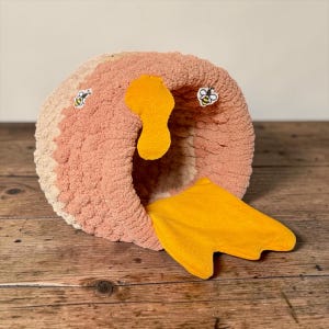 May include: A round, textured, crocheted pet cave in shades of dusty pink and cream. It features a yellow felt cutout shaped like a honeycomb hanging from the top and a yellow felt mat extending from the base. Two small bee patches are attached.