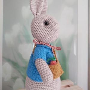 May include: A crocheted bunny rabbit wearing a blue shirt and carrying a basket with carrots. The bunny has a red and white checked ribbon around its neck.