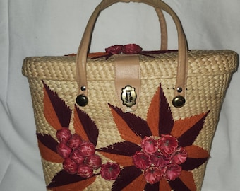 Könnte beinhalten: Eine gewebte Strohhandtasche mit rechteckiger Form und einem oberen Griff. Die Tasche hat ein Blumendesign mit roten und orangefarbenen Stickakzenten. Sie hat einen Lederriemenverschluss und goldfarbene Hardware.