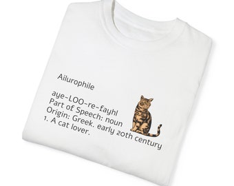 Pode incluir: Camiseta branca com a palavra "Ailurophile" e sua definição, acompanhada por um gráfico de um gato tigrado. A definição inclui a pronúncia, a parte do discurso, a origem e o significado: "Amante de gatos."