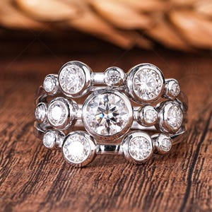 May include: A silver ring set with multiple clear, round gemstones. The ring has a cluster design with various sized stones, creating a sparkling effect. The ring is displayed on a wooden surface, highlighting the jewellery's brilliance.