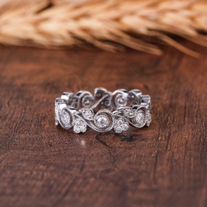 May include: A silver-toned eternity band featuring a continuous pattern of swirling metal vines and leaves. The ring is set with multiple small, round, clear stones embedded within the metal filigree, creating a textured and detailed surface.