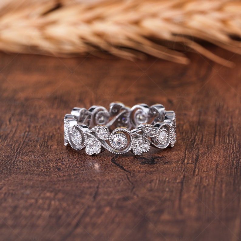 May include: A silver-toned eternity band featuring a continuous pattern of swirling metal vines and leaves. The ring is set with multiple small, round, clear stones embedded within the metal filigree, creating a textured and detailed surface.