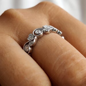 May include: A silver-toned metal ring featuring a series of small, round, clear stones set within a decorative, swirling filigree pattern. The band displays intricate milgrain detailing along the edges, creating a textured, vintage-inspired appearance.