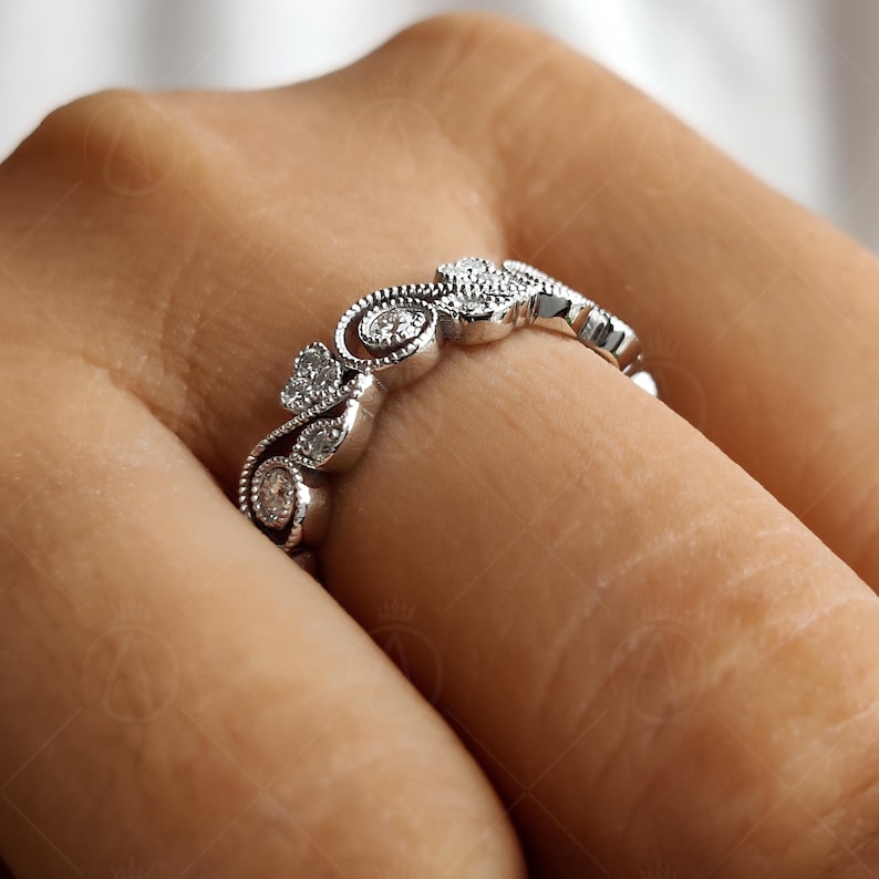 May include: A silver-toned metal ring featuring a series of small, round, clear stones set within a decorative, swirling filigree pattern. The band displays intricate milgrain detailing along the edges, creating a textured, vintage-inspired appearance.