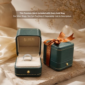 May include: A dark green square jewelry box with gold stitching and a gold button clasp. One box is open, revealing a silver ring with embedded stones on a beige velvet insert, with the Asuri Jewels logo printed inside. A second box is closed with a brown ribbon.