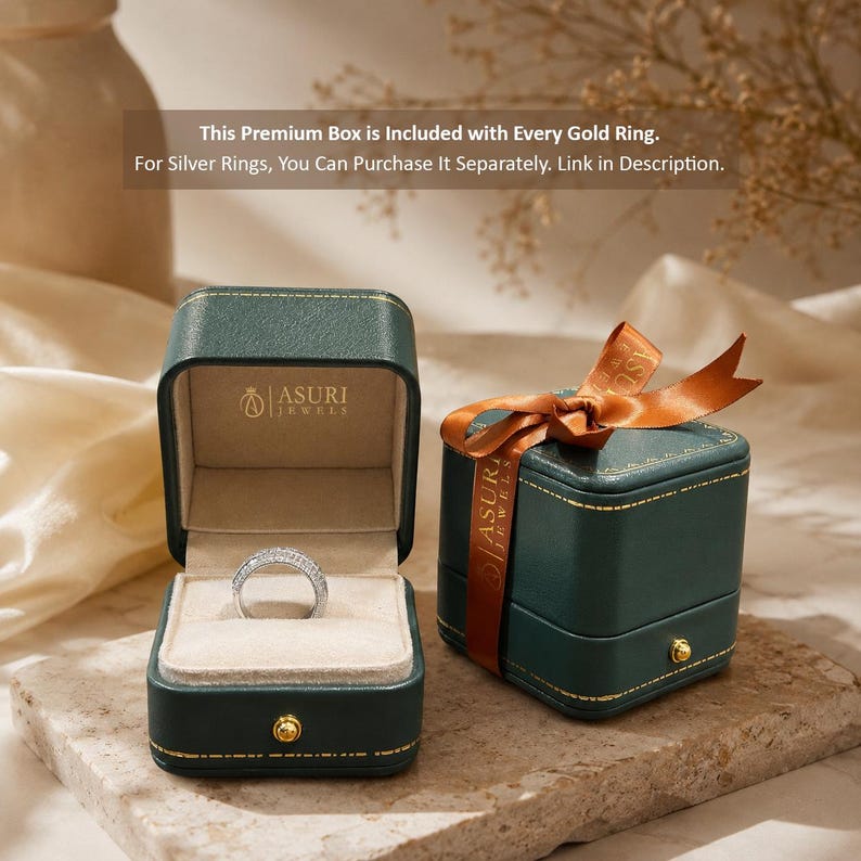 May include: A dark green square jewelry box with gold stitching and a gold button clasp. One box is open, revealing a silver ring with embedded stones on a beige velvet insert, with the Asuri Jewels logo printed inside. A second box is closed with a brown ribbon.