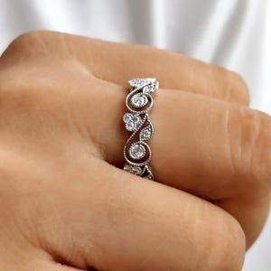 May include: A silver-toned ring featuring an intricate, openwork band design with swirling metal patterns and small, round, clear stones embedded within the filigree. The metalwork has a textured, beaded edge detail along the band's curves.