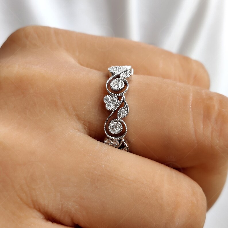 May include: A silver-toned ring featuring an intricate, openwork band design with swirling metal patterns and small, round, clear stones embedded within the filigree. The metalwork has a textured, beaded edge detail along the band's curves.