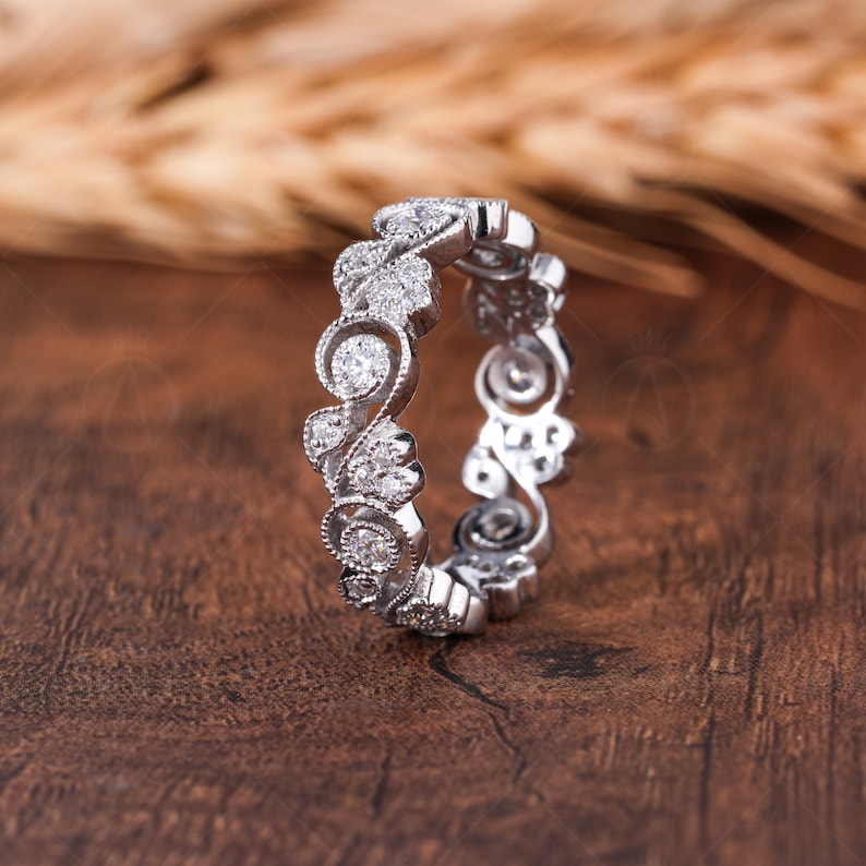 May include: A silver-toned eternity band featuring a continuous pattern of swirling metal filigree and small, round, clear stones set throughout the design. The ring has a textured, beaded edge along the metalwork, creating a detailed, intricate appearance.