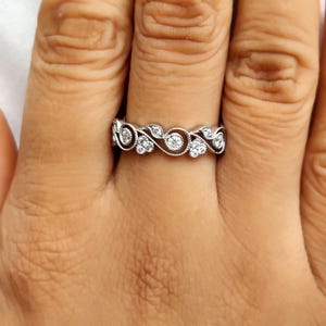 May include: A silver-toned metal ring featuring a continuous band with an intricate swirling vine pattern. The design is accented with small, round, clear stones set within the metal curves, creating a textured and detailed appearance on the band.