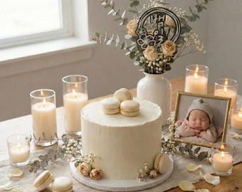 May include: A white frosted cake topped with macarons, surrounded by lit candles, flowers, and a framed baby photo. The cake is the centerpiece of a celebratory table setting, with a vase of flowers and a decorative topper.