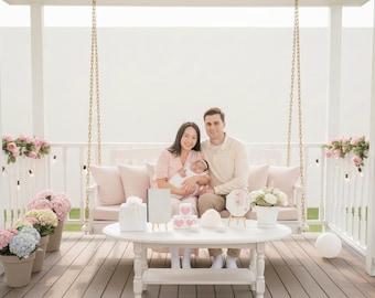 May include: A family poses on a white swing with pink cushions, surrounded by flowers and decorations. A round white table holds gifts and a cake. The scene is set on a wooden deck with a white pergola.