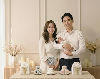 May include: A family portrait featuring a mother, father, and newborn baby. The parents are dressed in white, and the baby is held by the father. Decorative items, including a cake and gifts, are on a table in front of them.