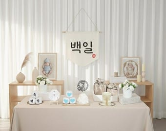 May include: A beige table is set with decorations for a baby's 100th-day celebration. The scene includes framed photos, gifts, flowers, and a banner with Korean text. The background features white curtains.