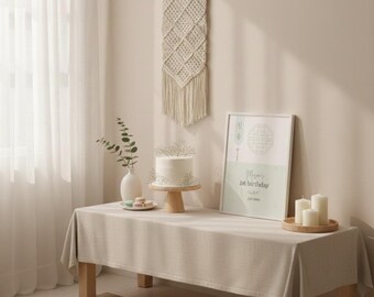 May include: A beige linen tablecloth covers a wooden table, set with a white cake on a wooden stand, a framed print with the text "Mom's 1st birthday", and candles. A macrame wall hanging and a vase with greenery are also visible.