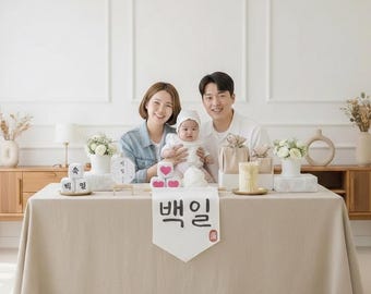 May include: A family of three smiles at the camera, gathered around a table set for a celebration. The table is covered with a beige tablecloth and decorated with gifts, flowers, and a banner with Korean text. A baby in a white outfit is held by the parents.
