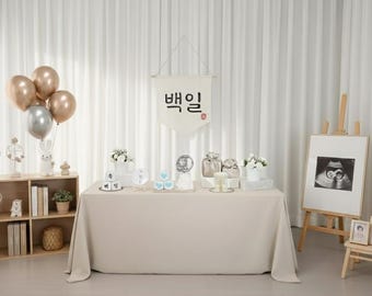 May include: A beige table is set for a celebration, adorned with balloons, gifts, and a banner with Korean text. A framed ultrasound image and a small wooden shelf with decor complete the scene. The backdrop is a white curtain.