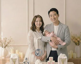 May include: A family portrait featuring a mother in a white lace dress, a father holding a baby, and a table with gifts and a cake. The cake has a sign with Korean text. The gifts are wrapped in white paper with gold butterfly accents.