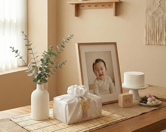 May include: A serene interior scene featuring a framed baby photo, a white cake on a stand, and a decorative white gift box with a gold butterfly accent. A vase with eucalyptus and a wooden block with Korean text are also present.