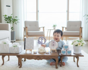 May include: A smiling baby sits near a low wooden table with traditional Korean first birthday items. The table holds wrapped gifts, a calligraphy board, a decorative fan, candles, and heart-shaped blocks. A vase of white flowers is also present.