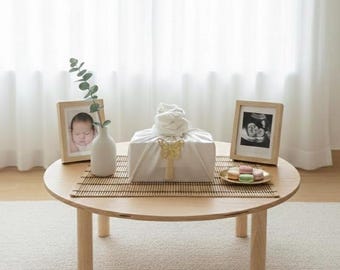 May include: A round wooden coffee table with a bamboo mat, holding a wrapped gift, framed baby photos, a vase with greenery, and a plate of macarons. The gift is white with a gold bow. The room has white curtains.
