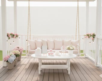 May include: A white swing bench with pink cushions hangs from a white pergola. A white oval table holds decorative items, including flowers, hearts, and a gift bag. Potted hydrangeas in pink, blue, and yellow sit nearby.